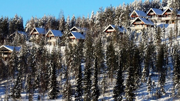 Une rangée de cabanes en bois aux toits couverts de neige, entourées d ’épais conifères, par une journée ensoleillée.