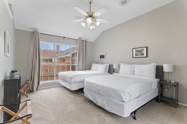 A bedroom with two beds, a ceiling fan, a window with curtains, and a small bedside table with a lamp.
