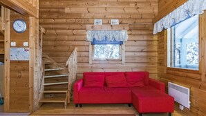 Interior de una cabaña de madera con un sofá rojo, paredes de madera, una escalera, un reloj y una ventana con vista a árboles.