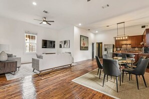 Amplia sala de estar con un sofá blanco, comedor con mesa y sillas de vidrio, y cocina moderna con gabinetes de madera y una lámpara colgante.