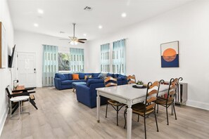 Ein modernes Wohnzimmer mit einem blauen Sofa, einem Esstisch mit Stühlen, einem Deckenventilator und einem Bild an der Wand.