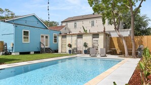 Ein Swimmingpool mit einer umlaufenden Terrasse, ein blaues Haus und ein weißes Haus im Hintergrund.