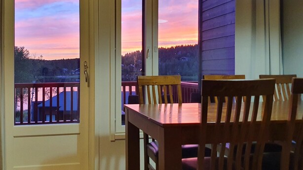 A dining area with a table and chairs, large windows with a view of a sunset over a forested landscape. - Skivillas 47 ukkohalla (a1) by Interhome (Hyrynsalmi)