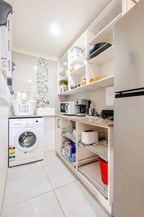 A compact kitchen with a washing machine, microwave, and shelves with various items. - The Wharf House @ Sussex Inlet (Sussex Inlet)