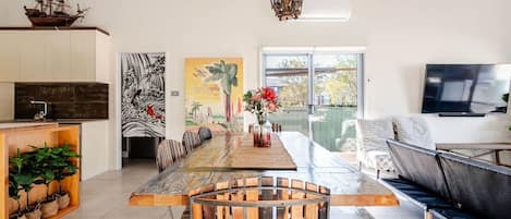 A spacious living area with a long wooden dining table, a comfortable sofa, and a television mounted on the wall.