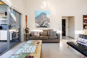 A living room with a sofa, a coffee table with a board game, a painting of a tree, and a bookshelf. - The Wharf House @ Sussex Inlet (Sussex Inlet)