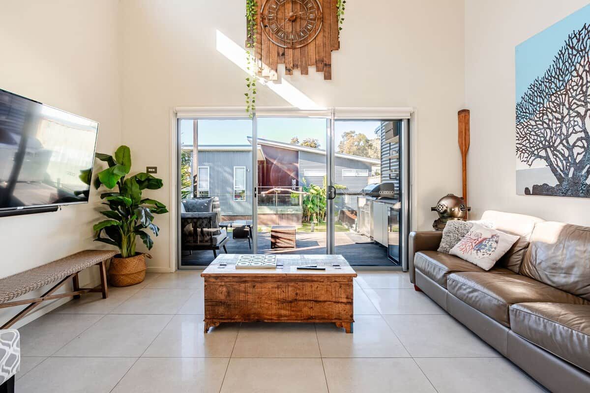 A modern living room with a flat-screen TV, a wooden coffee table, a leather sofa, a large window, and a view of the outdoors.