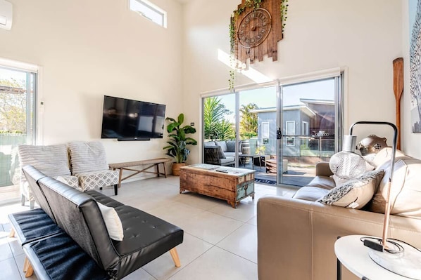 A modern living room with a flat-screen TV, leather sofas, a coffee table, and a large window offering a view of the outdoors.
