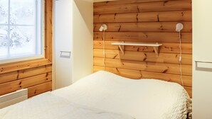 A bedroom with a wooden headboard, a bed with a white duvet, a window with blinds, and a shelf with a lamp.