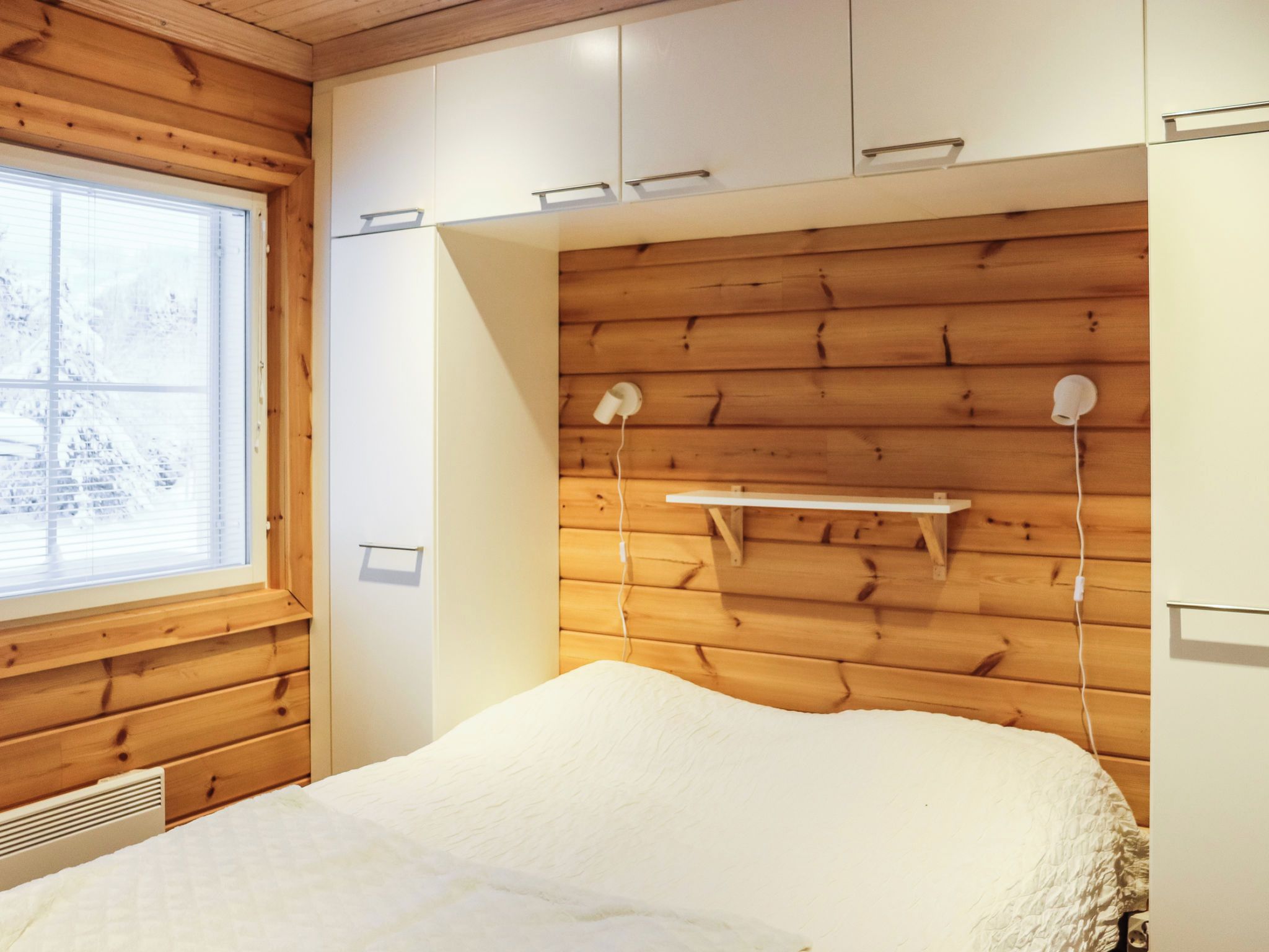 Une chambre avec une tête de lit en bois, un lit recouvert d’une couverture blanche, une fenêtre avec des stores et une étagère fixée au mur.