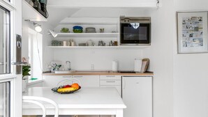 Een kleine keuken met een wit aanrecht, een kleine koelkast en een magnetron erboven. Op het aanrecht staat een schaal met fruit en een stoel met een gestreept kussen.