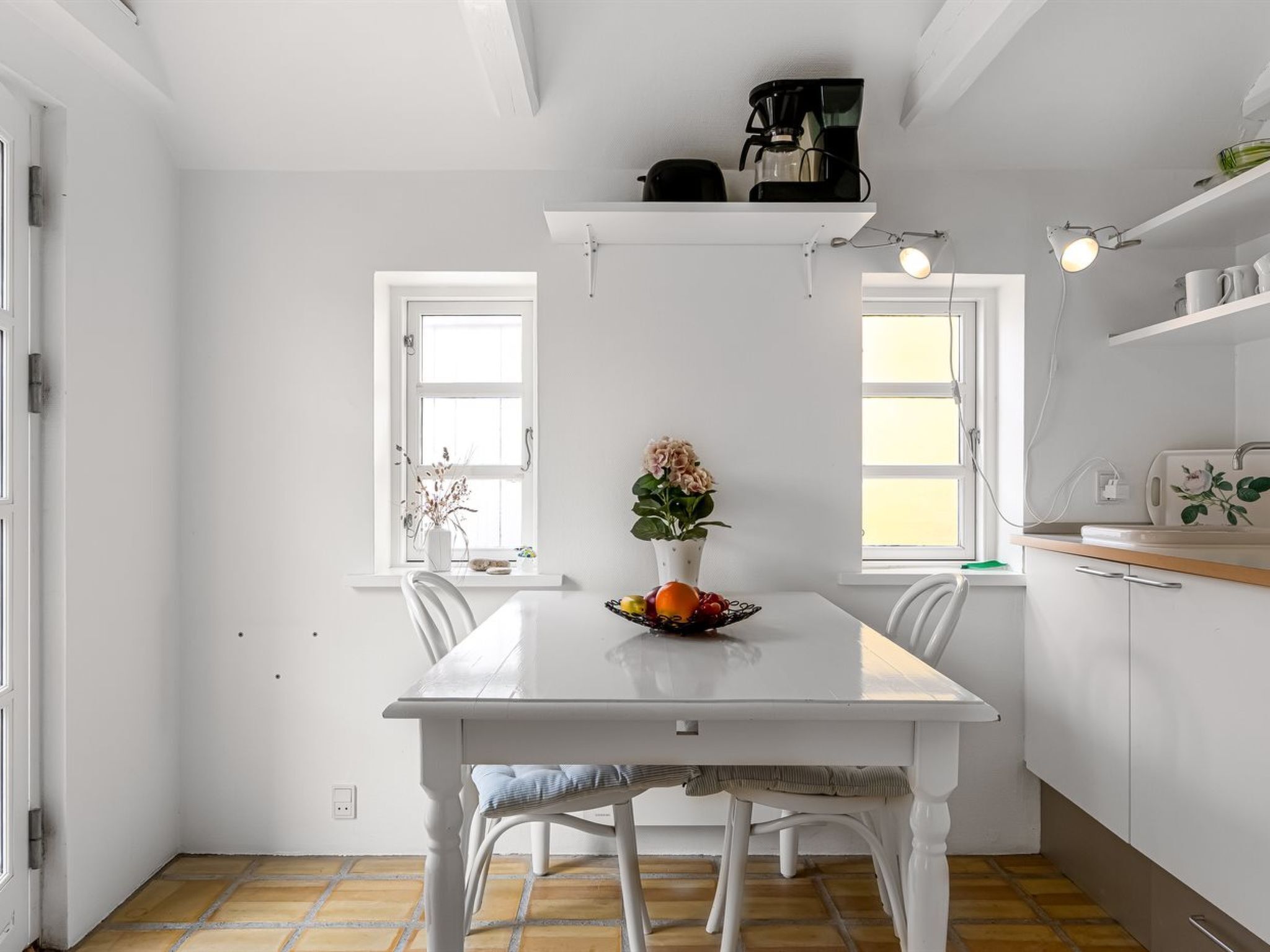 Una mesa de comedor blanca con sillas, un jarrón con flores y un frutero. Hay dos ventanas con marcos blancos y una estantería sobre la mesa que contiene una cafetera y una lámpara.