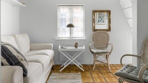 Een gezellige kamer met een witte bank, een klein tafeltje met een lamp, twee rieten stoelen, een kamerkleed met een patroon en een raam met een witte omlijsting.
