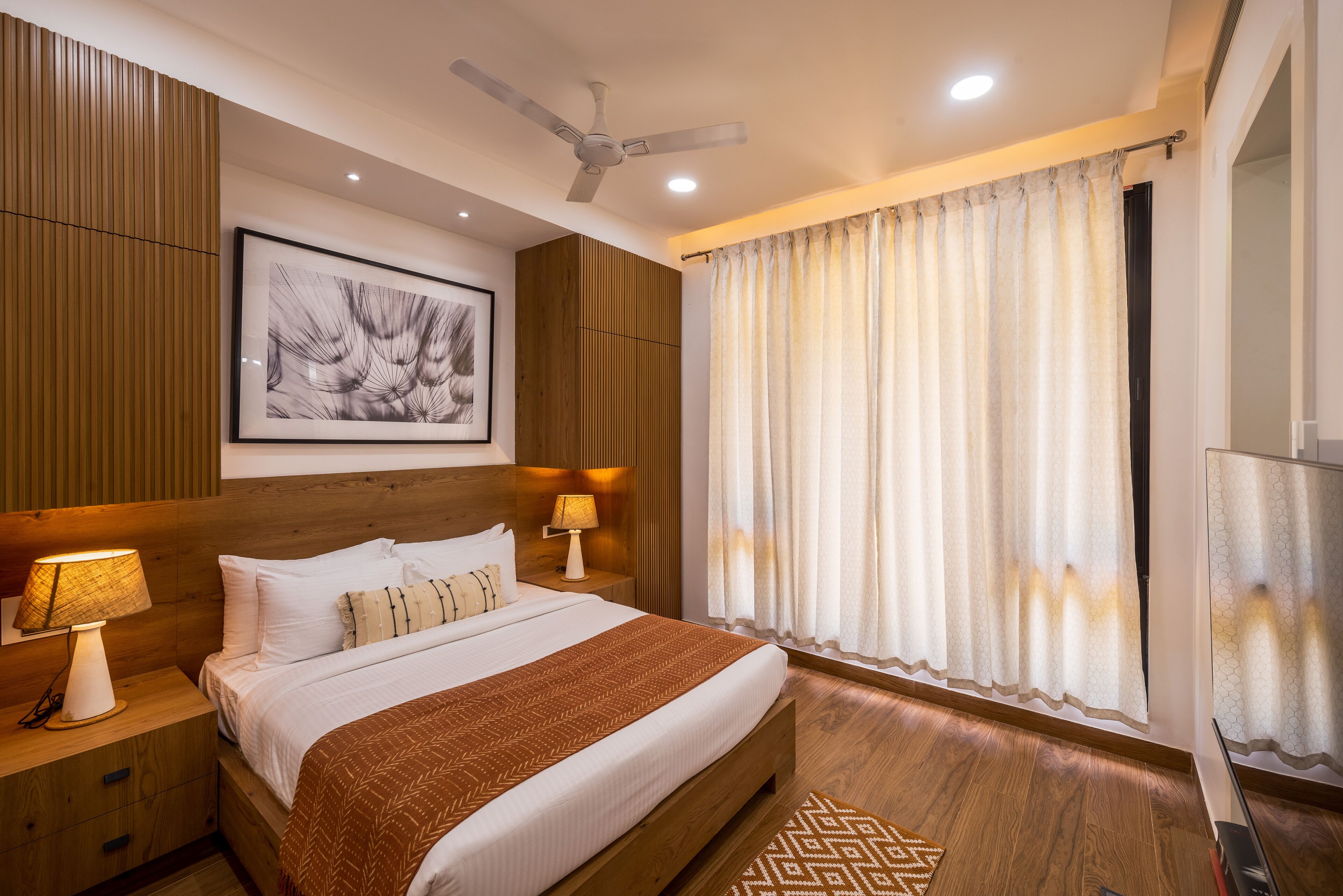A well-lit bedroom with a large bed, wooden paneling, a ceiling fan, and a framed artwork on the wall.
