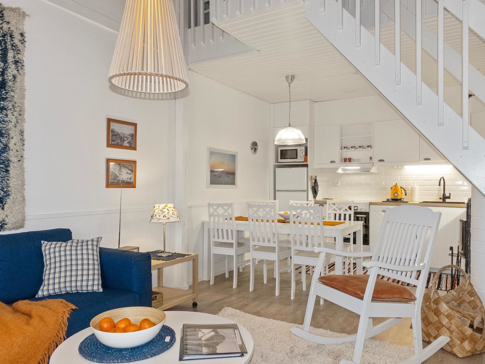 A modern living room with a blue sofa, a dining area with white chairs, a kitchen with white cabinets, and a staircase.