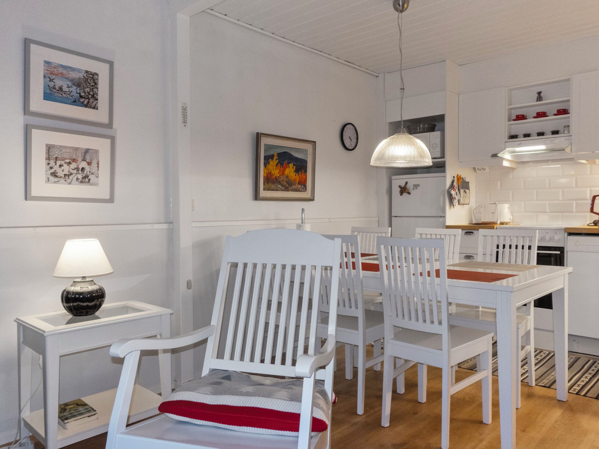 Een eethoek met witte stoelen en een tafel, een keuken met witte kastjes, en een wand met ingelijste kunstwerken.