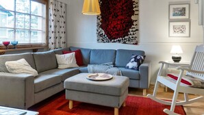 A modern living room with a grey sectional sofa, a small ottoman, a rocking chair, and a large abstract tapestry on the wall. - Päivänsäde by Interhome (Inari)