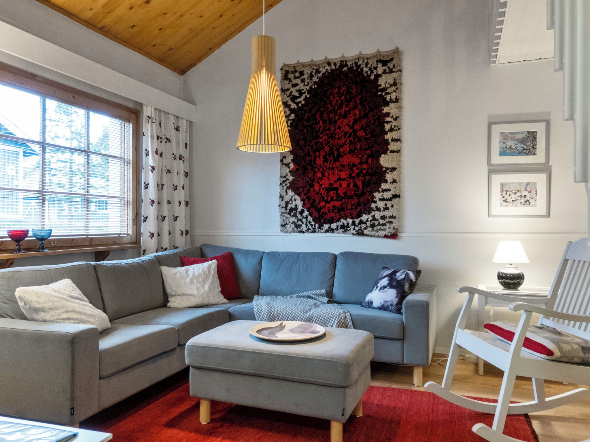 A modern living room with a grey sectional sofa, a red rug, a wooden ceiling, and a large abstract wall hanging.