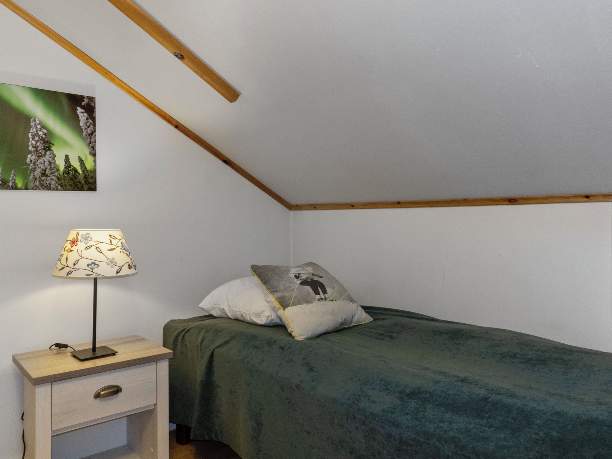 A small, simple room with a bed, a nightstand, a lamp, and a picture of the aurora borealis on the wall.