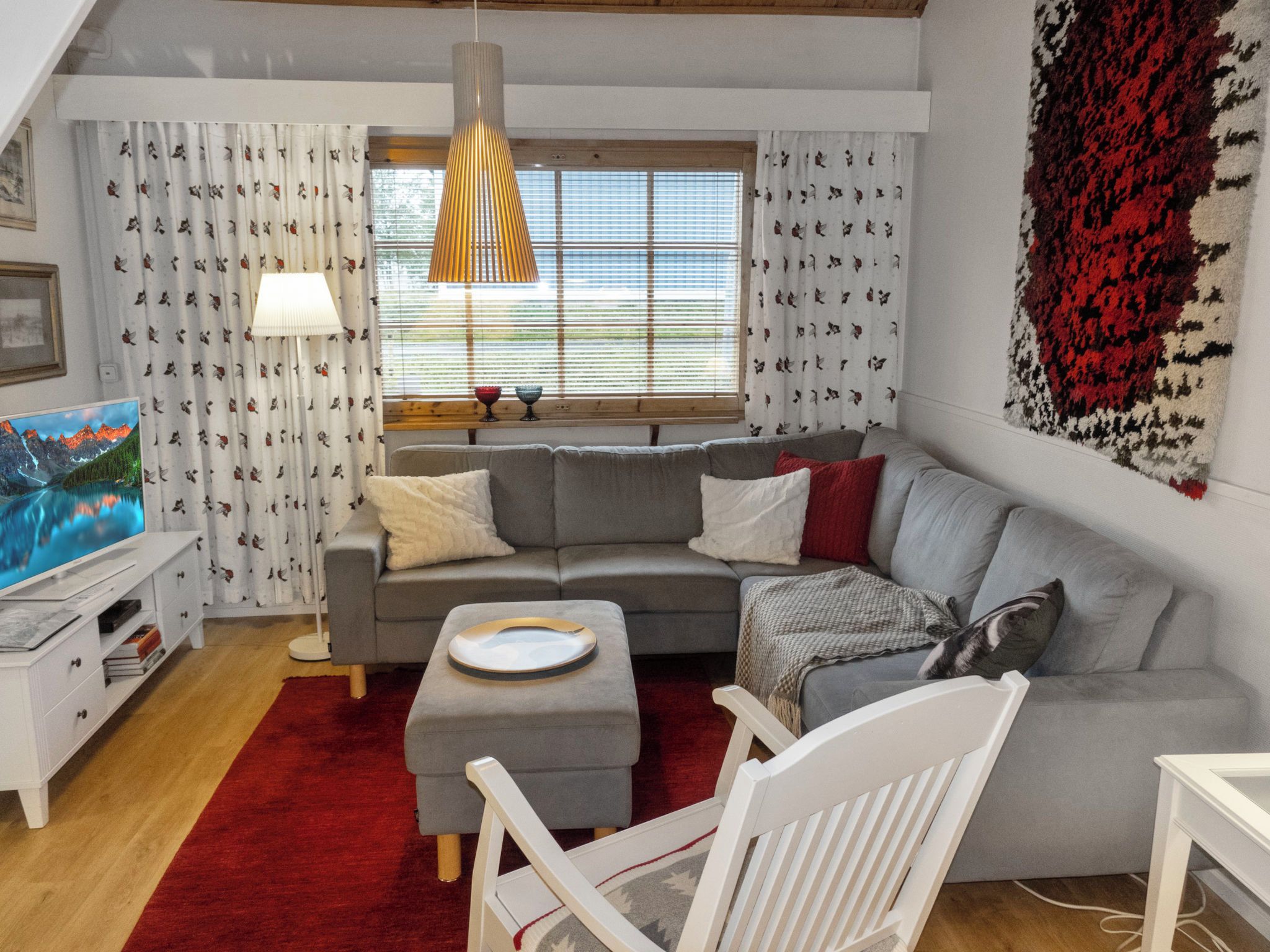 A modern living room with a grey sofa, a white armchair, a red rug, and a television displaying a mountain scene.