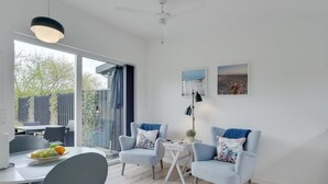 A modern living room with a dining area, featuring light blue armchairs, a white coffee table, and a large window offering a view of the outdoors.