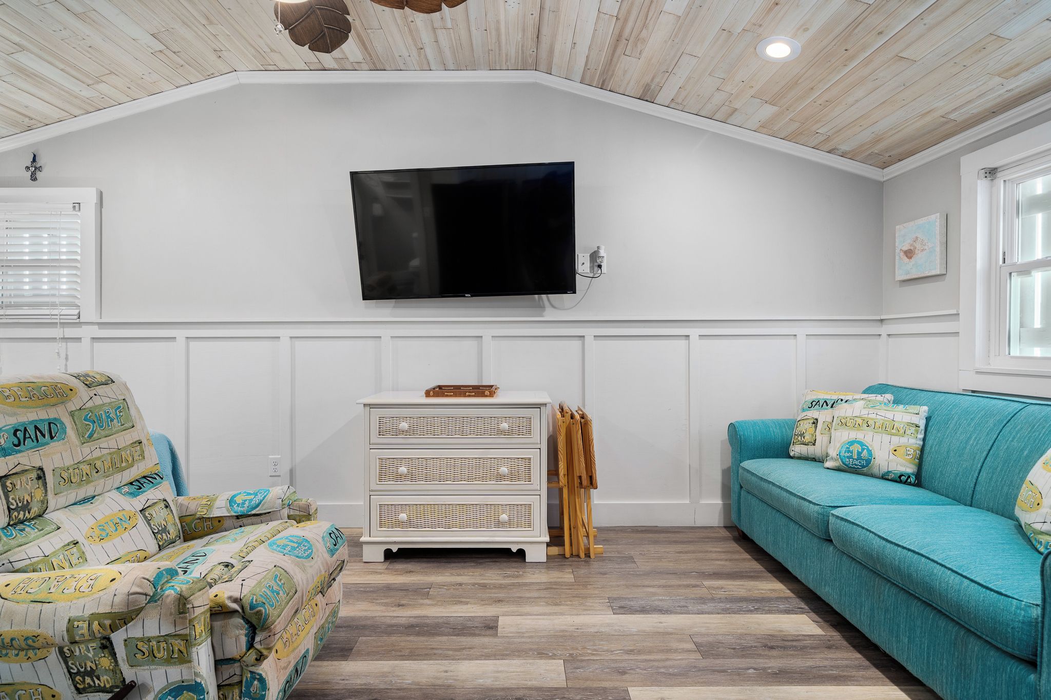 Una habitación con pared blanca, televisor montado, cómoda blanca, sofá color turquesa y sillón estampado.