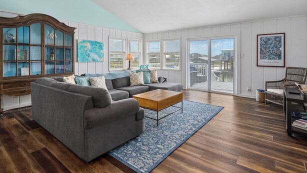 A spacious living room with a gray sectional sofa, a wooden coffee table, and a large window offering a view of the outside. - Hideaway at Redhead Road by Miss Kitty’s (Rockport)