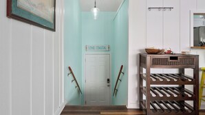 A hallway with a light blue wall, a wooden staircase, a door labeled "GONE COASTAL", and a wooden rack with shoes. - Hideaway at Redhead Road by Miss Kitty’s (Rockport)