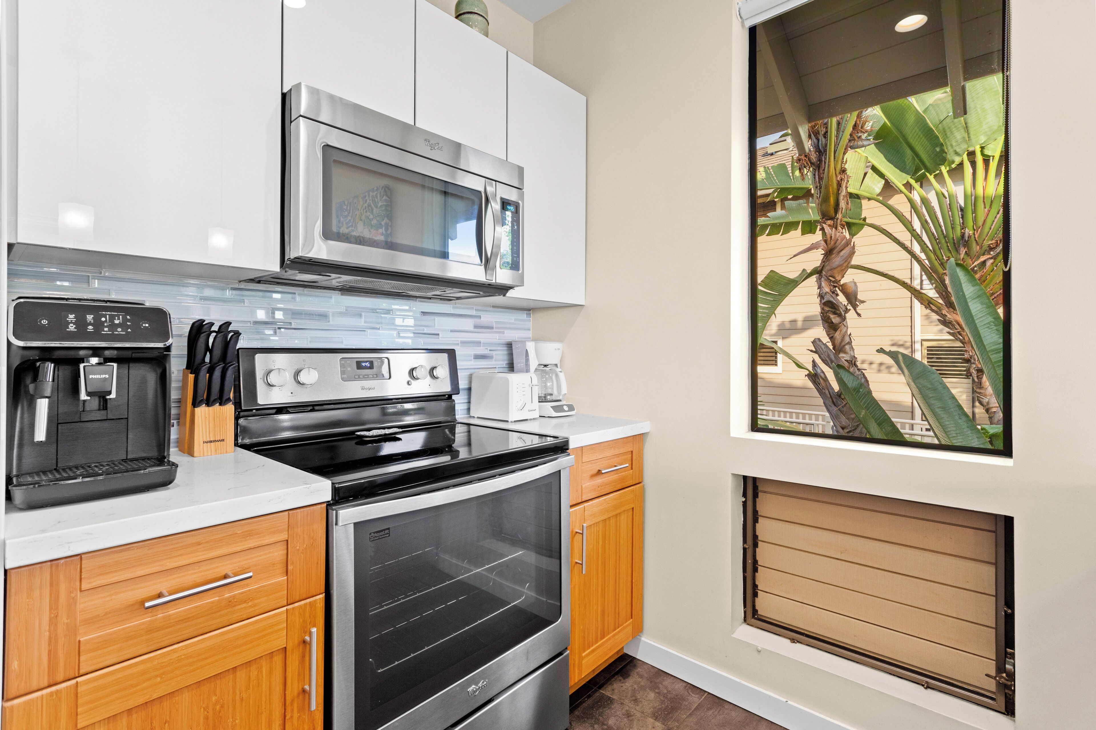 A modern kitchen with stainless steel appliances, wooden cabinets, and a view of a palm tree outside.