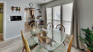 A dining area with a glass table and wicker chairs, a kitchen in the background, and a plant on the right. - A Bay From It All by Miss Kitty’s (Rockport)