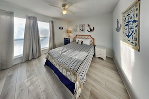 A bedroom with a bed, curtains, a ceiling fan, a nightstand, a lamp, and wall decorations.
