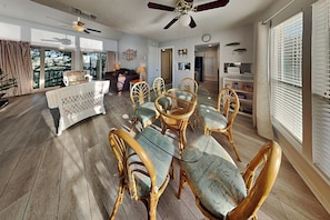 A spacious dining area with a glass-top table and wicker chairs, a ceiling fan, and a view through large windows.