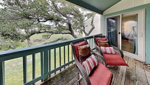 A covered porch with wicker chairs and red cushions, a wooden deck, and a view of trees and grass. - A Bay From It All by Miss Kitty’s (Rockport)
