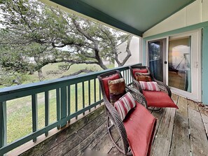 A covered porch with wicker chairs and red cushions, a wooden deck, and a view of a tree and lawn.