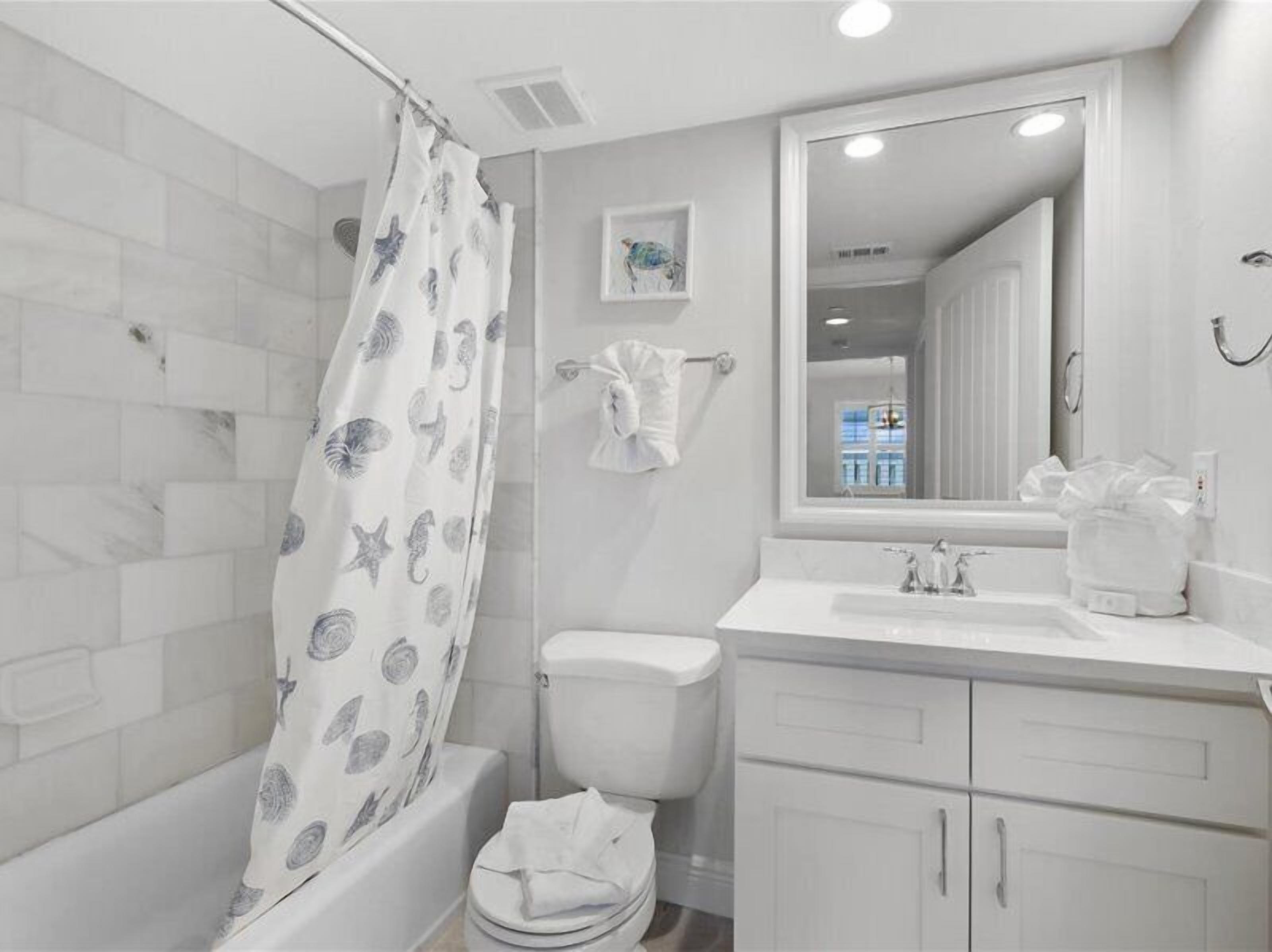 A modern bathroom with a shower curtain featuring sea creatures, a toilet, a sink with a mirror above it, and a small framed picture on the wall.