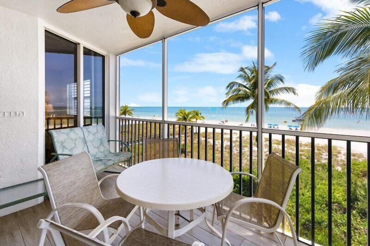 A balcony with a white table and chairs, a ceiling fan, and a view of the beach and palm trees.