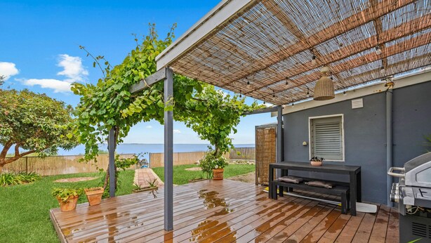 Eine Holzterrasse mit einer Pergola, die mit Grünpflanzen bewachsen ist, einem Grill und einer Sitzgelegenheit.