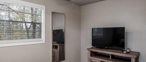 A bedroom with a ceiling fan, a TV on a wooden stand, a large mirror, and a window with blinds.