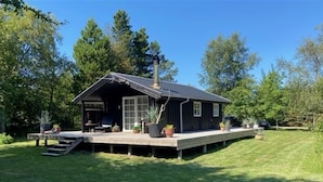 A small, dark-colored cabin with a covered porch, surrounded by a grassy yard and trees.