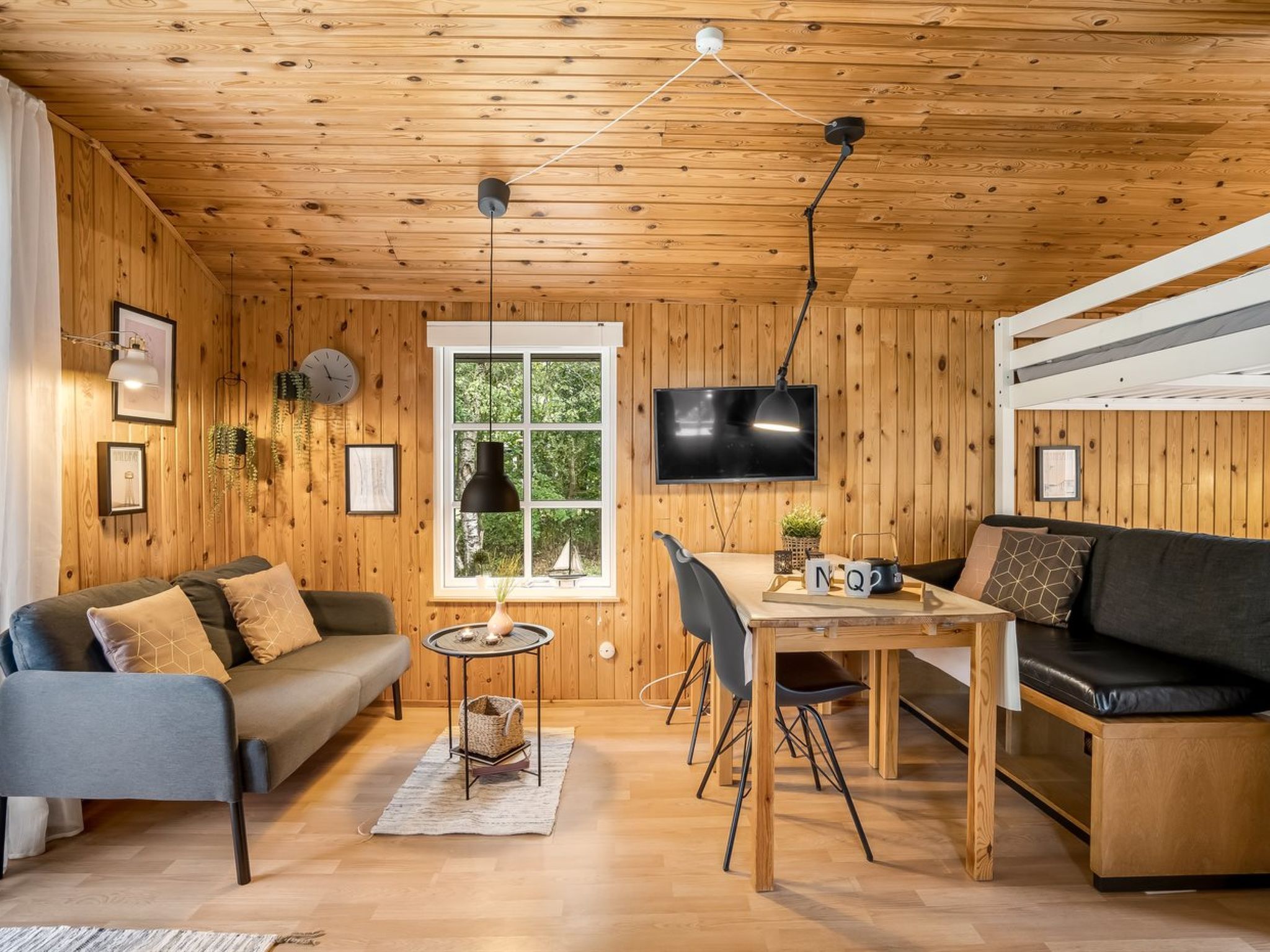 A cozy cabin interior with a wooden ceiling, a TV mounted on the wall, a sofa, a dining table with chairs, and a window with a view of greenery.