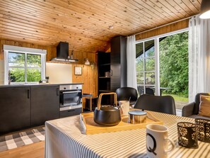 A cozy kitchen with wooden walls, a dining area with a table set for two, and large windows offering a view of greenery.