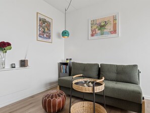 Uma sala de estar com um sofá verde, uma mesinha com velas, um cesto de vime, um banquinho decorativo e duas obras de arte emolduradas na parede.
