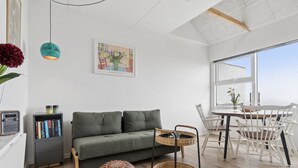 A modern living room with a sofa, a dining table with chairs, a bookshelf, a hanging light, and a large window.