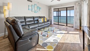 A modern living room with a leather sofa, a glass coffee table, and a view of the ocean through large sliding doors. - Arie Dam 404 by Ryson Vacation Rentals (Madeira Beach)