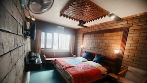 A room with a brick wall, a bed with a red and blue bedspread, a wooden chair, and a small table with a lamp. - Hotel Akbar  (Srinagar)