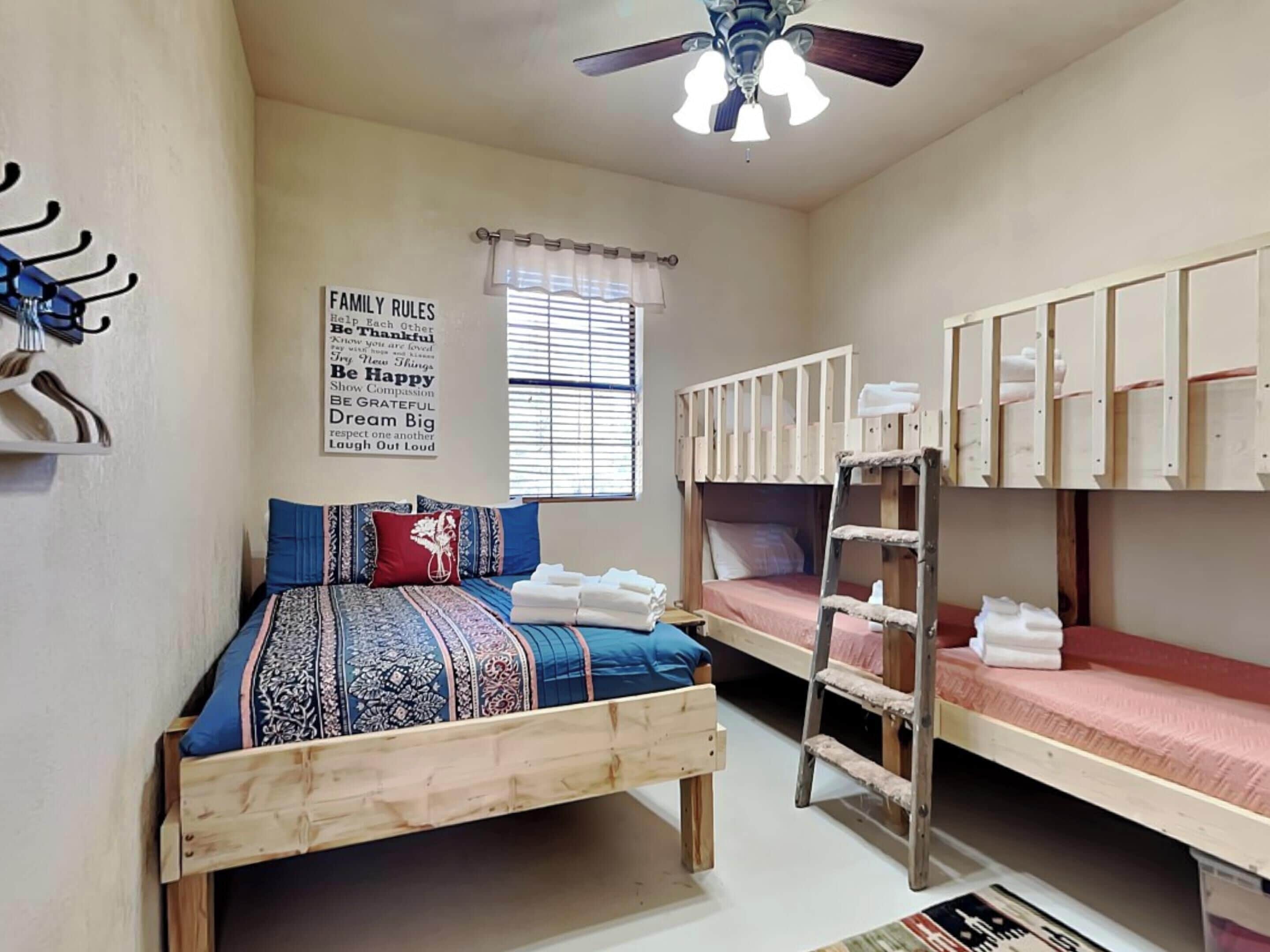A dormitory room with bunk beds, a ceiling fan, a window with curtains, and a wall sign with family rules.