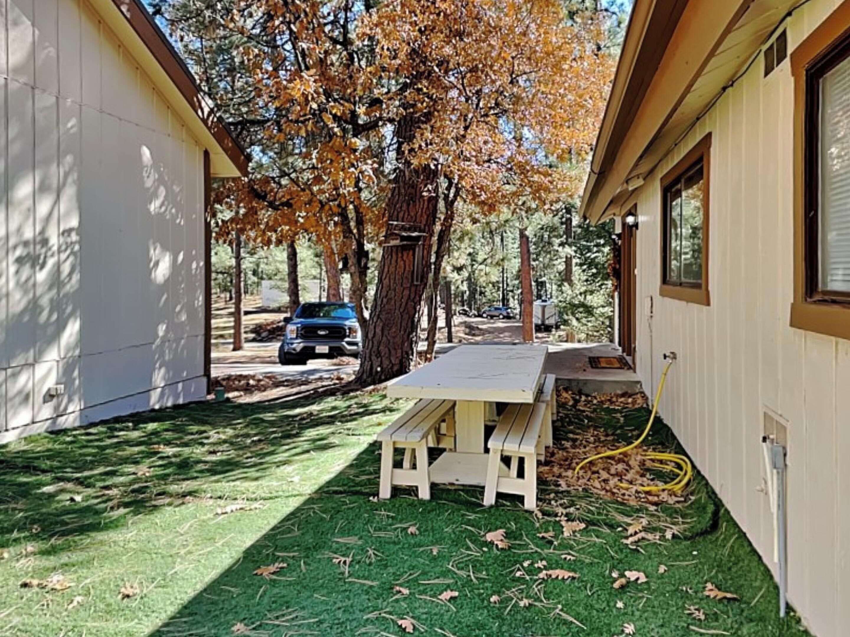 A covered patio with a picnic table, surrounded by artificial grass and trees with autumn leaves.
