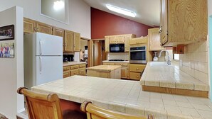 A kitchen with wooden cabinets, a white refrigerator, a microwave, an oven, and a sink. There are two wooden chairs at the island. - Quiet Mountain Hideaway by Ruidoso Vacation Rentals (Ruidoso)