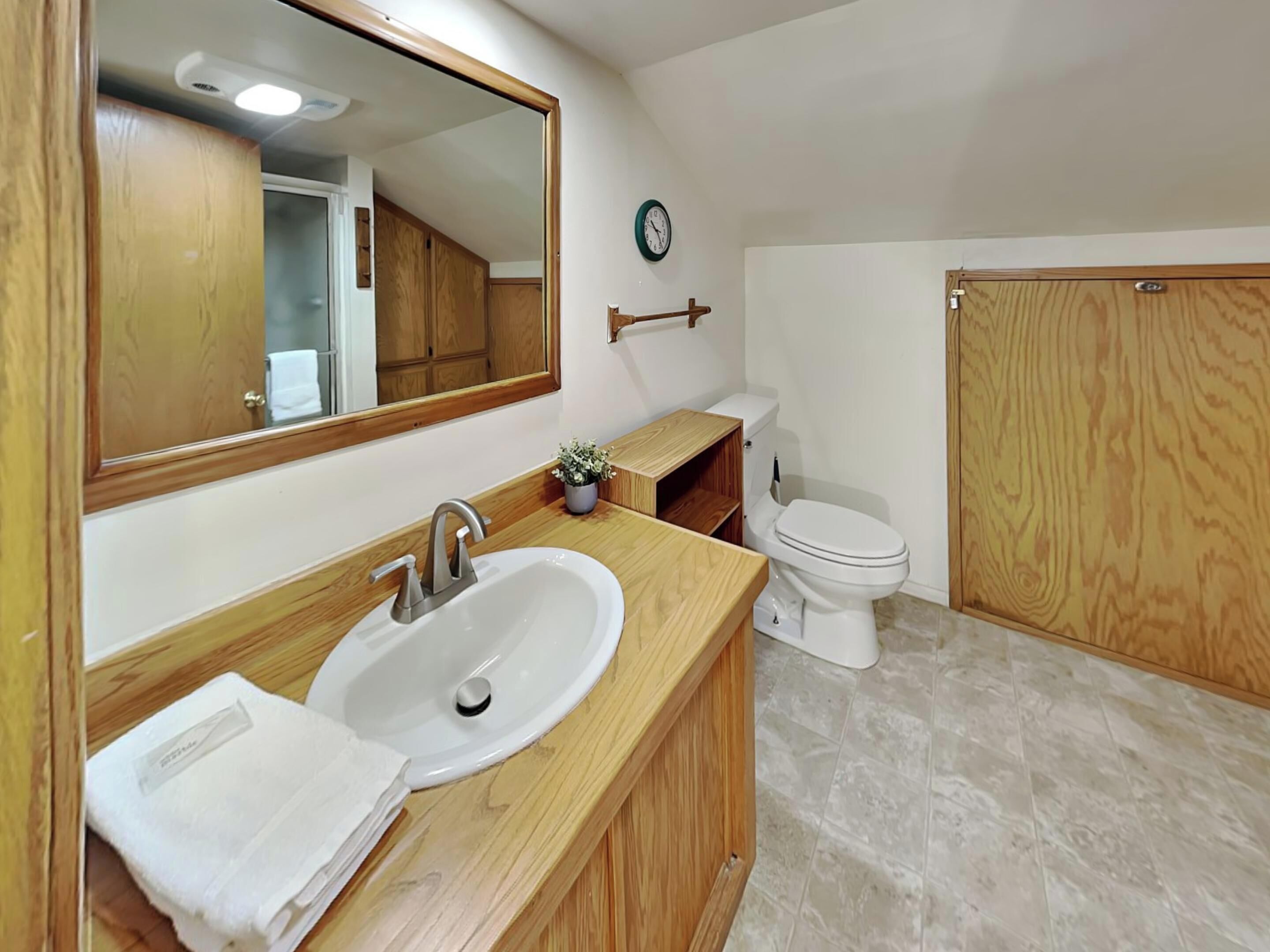 A bathroom with a wooden vanity, a sink, a mirror, a toilet, and a clock on the wall.