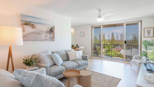 A modern living room with a sofa, a coffee table, a lamp, and a painting of a beach scene. - Poppy's Place - Oceanstays (Yamba)